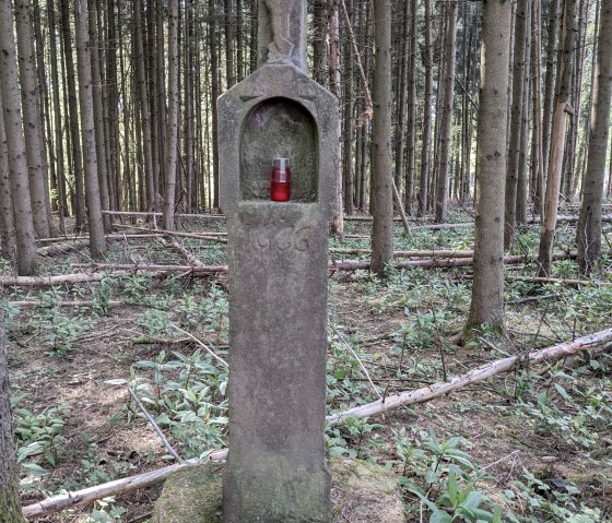 Een stenen wegkruis met een Christusfiguur en een rood graflicht staat in een dicht bos., © A. Girards Een stenen wegkruis met een Christusfiguur en een rood graflicht staat in een dicht bos., © A. Girards