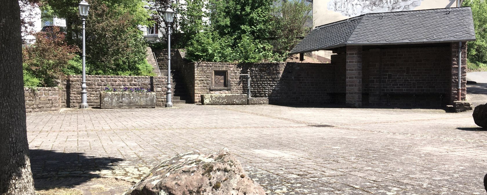 Een rustig dorpsplein met een geplaveide vloer, omringd door bomen en een gebouw met muurschilderingen. Lantaarns en planten sieren het plein., © Tourist-Information Bitburger Land Een rustig dorpsplein met een geplaveide vloer, omringd door bomen en een gebouw met muurschilderingen. Lantaarns en planten sieren het plein., © Tourist-Information Bitburger Land