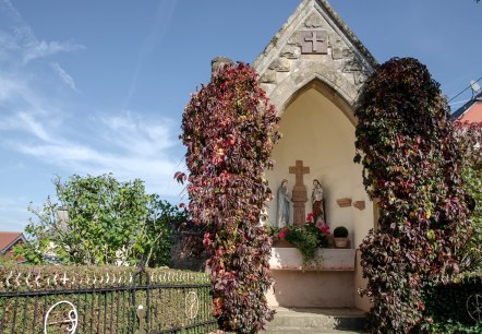 Une petite chapelle à Wolsfeld, entourée de plantes grimpantes rouges. A l'intérieur, on peut voir des statues religieuses. Le ciel est clair et bleu., © Tourist-Information Bitburger Land Une petite chapelle à Wolsfeld, entourée de plantes grimpantes rouges. A l'intérieur, on peut voir des statues religieuses. Le ciel est clair et bleu., © Tourist-Information Bitburger Land
