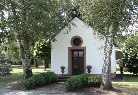 Eine kleine weiße Kapelle mit braunen Akzenten, umgeben von Bäumen und Sträuchern in einer grünen Landschaft., © TI Bitburger Land Eine kleine weiße Kapelle mit braunen Akzenten, umgeben von Bäumen und Sträuchern in einer grünen Landschaft., © TI Bitburger Land
