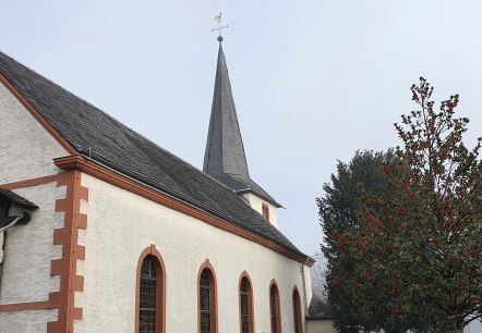 Eine Kirche in Idesheim mit einem Kreuz an der Fassade und einem Baum mit roten Beeren im Vordergrund., © TI Bitburger Land Eine Kirche in Idesheim mit einem Kreuz an der Fassade und einem Baum mit roten Beeren im Vordergrund., © TI Bitburger Land