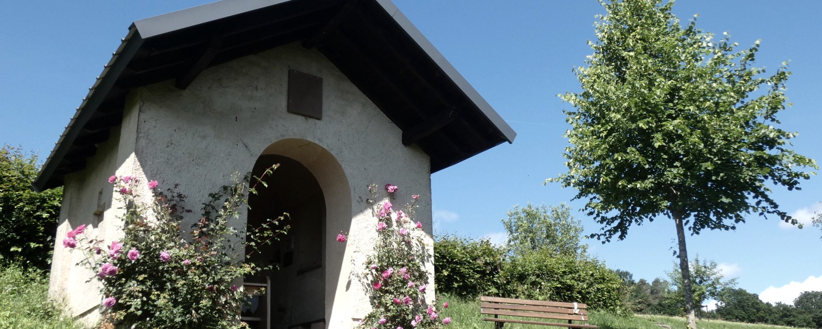 Petite chapelle dans la verdure, entourée de roses en fleurs. Un banc se trouve à côté, un arbre fait de l'ombre. Ciel bleu en arrière-plan., © Tourist-Information Bitburger Land, Melanie Salzburger Petite chapelle dans la verdure, entourée de roses en fleurs. Un banc se trouve à côté, un arbre fait de l'ombre. Ciel bleu en arrière-plan., © Tourist-Information Bitburger Land, Melanie Salzburger