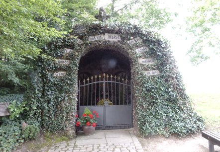 Eine mit Efeu bewachsene Lourdesgrotte mit einem Tor. Auf dem Tor sind Blumen zu sehen. Schilder mit Schriftzügen zieren den Eingang., © Eifelverein Ortsgruppe Speicher Eine mit Efeu bewachsene Lourdesgrotte mit einem Tor. Auf dem Tor sind Blumen zu sehen. Schilder mit Schriftzügen zieren den Eingang., © Eifelverein Ortsgruppe Speicher