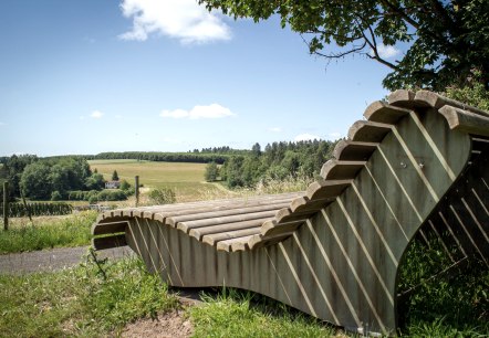 Banc de repos en bois avec vue sur des champs et des forêts verdoyants sous un ciel bleu à Neidenbach., © TI Bitburger Land - Monika Mayer Banc de repos en bois avec vue sur des champs et des forêts verdoyants sous un ciel bleu à Neidenbach., © TI Bitburger Land - Monika Mayer