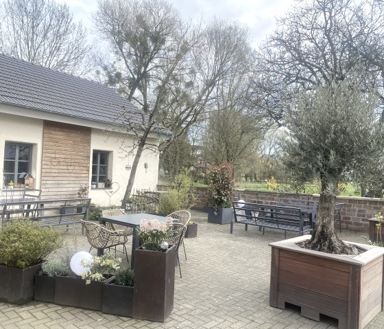 Ein gemütlicher Außenbereich eines Cafés mit Tischen, Stühlen und Pflanzen. Im Hintergrund ein Gebäude mit Holzelementen und Bäumen., © TI Bitburger Land Ein gemütlicher Außenbereich eines Cafés mit Tischen, Stühlen und Pflanzen. Im Hintergrund ein Gebäude mit Holzelementen und Bäumen., © TI Bitburger Land