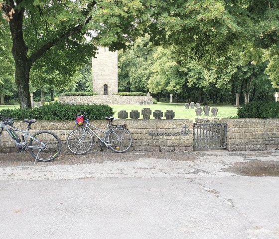 Deux vélos sont adossés à un mur de pierre. Derrière, un cimetière avec des pierres tombales et une tour, entouré d'arbres., © TI Bitburger Land Deux vélos sont adossés à un mur de pierre. Derrière, un cimetière avec des pierres tombales et une tour, entouré d'arbres., © TI Bitburger Land