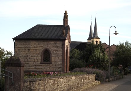Steinkapelle mit spitzem Dach und buntem Fenster, umgeben von Blumen. Im Hintergrund sind Kirchtürme zu sehen., © Thomas Neises Steinkapelle mit spitzem Dach und buntem Fenster, umgeben von Blumen. Im Hintergrund sind Kirchtürme zu sehen., © Thomas Neises