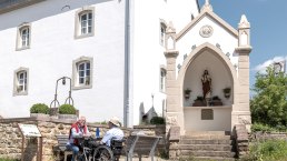 Zwei Männer, einer im Rollstuhl, sitzen auf einer Bank vor einem weißen Gebäude mit einer religiösen Statue in einer Nische. Sonniger Tag., © Naturpark Südeifel, Thomas Urbany Zwei Männer, einer im Rollstuhl, sitzen auf einer Bank vor einem weißen Gebäude mit einer religiösen Statue in einer Nische. Sonniger Tag., © Naturpark Südeifel, Thomas Urbany