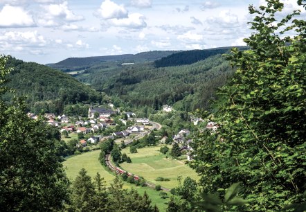 Blick auf St. Thomas mit grünen Hügeln, einem Dorf im Tal und einer Bahnstrecke. Der Himmel ist leicht bewölkt., © TI Bitburger Land Blick auf St. Thomas mit grünen Hügeln, einem Dorf im Tal und einer Bahnstrecke. Der Himmel ist leicht bewölkt., © TI Bitburger Land