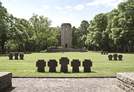 Un monument de guerre avec des croix en pierre sur une prairie, une tour en arrière-plan, entourée d'arbres verts sous un ciel bleu., © TI Bitburger Land Un monument de guerre avec des croix en pierre sur une prairie, une tour en arrière-plan, entourée d'arbres verts sous un ciel bleu., © TI Bitburger Land