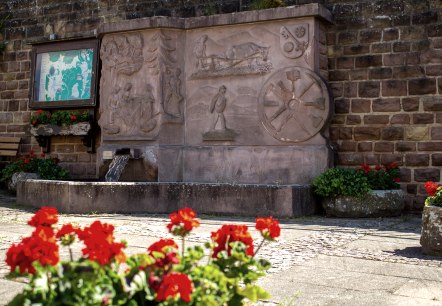 Une fontaine de village avec des reliefs artistiques en pierre, entourée de fleurs rouges. Un panneau d'information et un banc sont également visibles., © TI Bitburger Land M. Mayer Une fontaine de village avec des reliefs artistiques en pierre, entourée de fleurs rouges. Un panneau d'information et un banc sont également visibles., © TI Bitburger Land M. Mayer