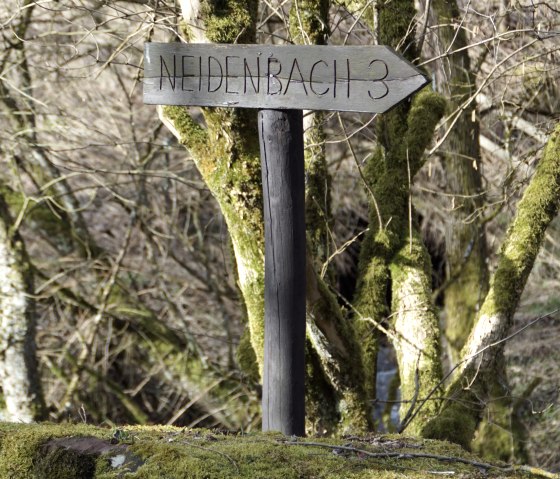 Ein hölzerner Wegweiser mit der Aufschrift 'Neidenbach 3' steht in einem moosbewachsenen Waldgebiet., © M. Bach Ein hölzerner Wegweiser mit der Aufschrift 'Neidenbach 3' steht in einem moosbewachsenen Waldgebiet., © M. Bach