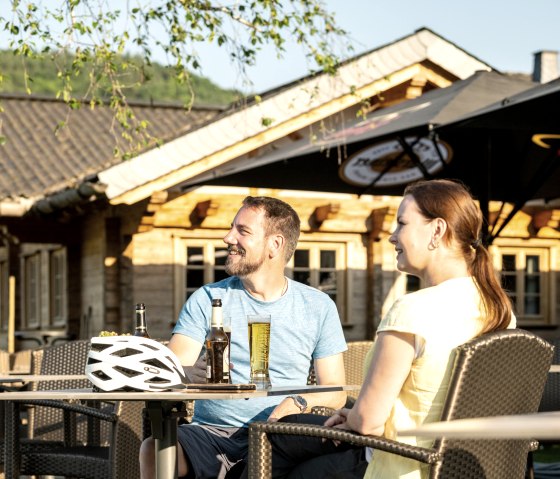 Zwei Personen sitzen entspannt an einem Tisch im Freien, genießen Getränke. Ein Fahrradhelm liegt auf dem Tisch. Im Hintergrund ein Holzgebäude., © Eifel Tourismus GmbH, Dominik Ketz Zwei Personen sitzen entspannt an einem Tisch im Freien, genießen Getränke. Ein Fahrradhelm liegt auf dem Tisch. Im Hintergrund ein Holzgebäude., © Eifel Tourismus GmbH, Dominik Ketz