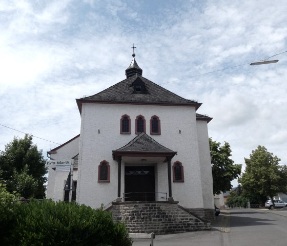 Witte kerk met rood raamkozijn, omringd door bomen en auto's. De lucht is bewolkt., © TI Bitburger Land Witte kerk met rood raamkozijn, omringd door bomen en auto's. De lucht is bewolkt., © TI Bitburger Land