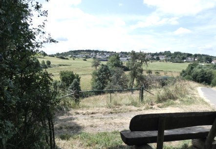 Eine Holzbank steht am Rand eines Feldwegs mit Blick auf eine grüne Landschaft und ein Dorf in der Ferne unter blauem Himmel., © TI Bitburger Land Eine Holzbank steht am Rand eines Feldwegs mit Blick auf eine grüne Landschaft und ein Dorf in der Ferne unter blauem Himmel., © TI Bitburger Land