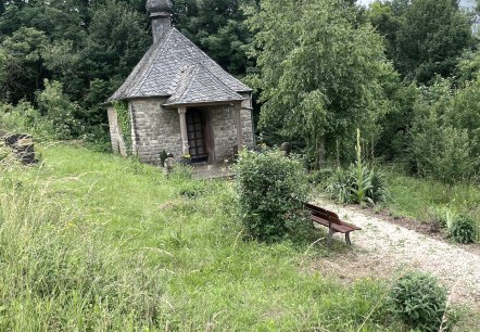 Eine kleine Steinkapelle mit Schieferdach steht inmitten üppiger Vegetation. Ein Kiesweg führt zu einer Holzbank vor der Kapelle., © Daniel Köhler Eine kleine Steinkapelle mit Schieferdach steht inmitten üppiger Vegetation. Ein Kiesweg führt zu einer Holzbank vor der Kapelle., © Daniel Köhler