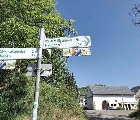 Wegwijzer toont afstanden naar Echternacherbrück, Minden, Wasserbilligerbrück en Ralingen. Een gebouw op de achtergrond, blauwe lucht., © Tourist-Information Bitburger Land, Steffi Wagner Wegwijzer toont afstanden naar Echternacherbrück, Minden, Wasserbilligerbrück en Ralingen. Een gebouw op de achtergrond, blauwe lucht., © Tourist-Information Bitburger Land, Steffi Wagner