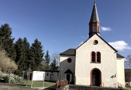Eine Kirche in Wißmannsdorf mit spitzem Turm und roten Fensterrahmen vor einem klaren blauen Himmel., © TI Bitburger Land Eine Kirche in Wißmannsdorf mit spitzem Turm und roten Fensterrahmen vor einem klaren blauen Himmel., © TI Bitburger Land