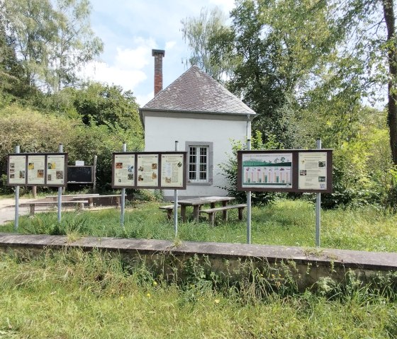 Un petit bâtiment blanc avec des panneaux d'information et des bancs devant, entouré d'arbres et d'herbe., © TI Bitburger Land Un petit bâtiment blanc avec des panneaux d'information et des bancs devant, entouré d'arbres et d'herbe., © TI Bitburger Land