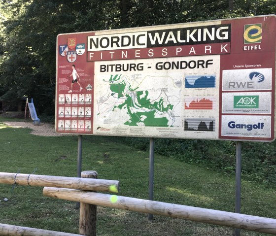 Schild des Nordic Walking Fitnessparks Bitburg-Gondorf mit Karte, Streckenprofilen und Sponsorenlogos. Im Hintergrund ein Spielplatz mit Rutsche., © TI Bitburger Land Schild des Nordic Walking Fitnessparks Bitburg-Gondorf mit Karte, Streckenprofilen und Sponsorenlogos. Im Hintergrund ein Spielplatz mit Rutsche., © TI Bitburger Land