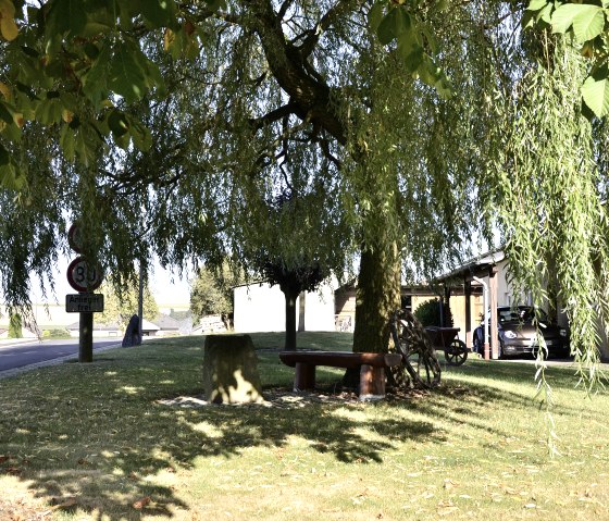 A shady rest area with a bench under a tree next to a road in Stockem, South Eifel Nature Park. A car is parked in the driveway of a house., © TI Bitburger Land A shady rest area with a bench under a tree next to a road in Stockem, South Eifel Nature Park. A car is parked in the driveway of a house., © TI Bitburger Land