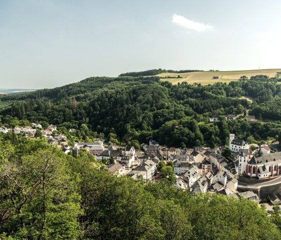 Panoramablick auf Neuerburg mit einer Kirche im Vordergrund, umgeben von dicht bewaldeten Hügeln und einem Windrad im Hintergrund., © Eifel Tourismus GmbH, D. Ketz Panoramablick auf Neuerburg mit einer Kirche im Vordergrund, umgeben von dicht bewaldeten Hügeln und einem Windrad im Hintergrund., © Eifel Tourismus GmbH, D. Ketz