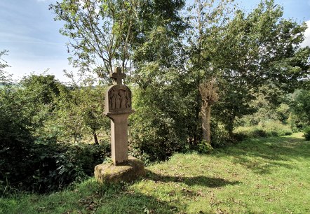 A stone cross stands on a green meadow, surrounded by trees and bushes. The sky is blue with a few clouds., © Tourist-Information Bitburger Land A stone cross stands on a green meadow, surrounded by trees and bushes. The sky is blue with a few clouds., © Tourist-Information Bitburger Land
