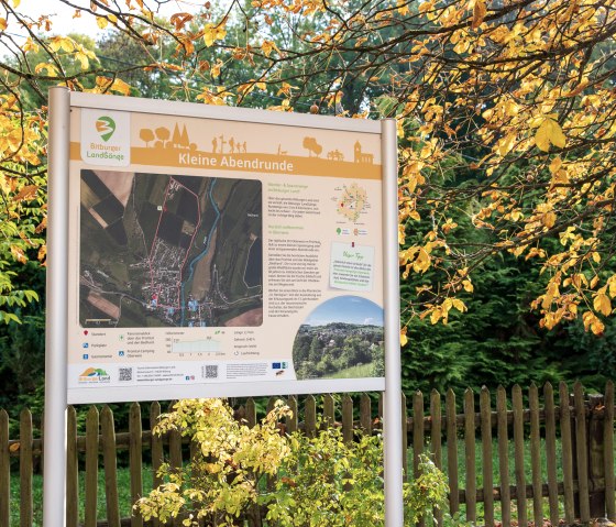 Bord voor de wandelroute 'Kleine Abendrunde' in Bitburger Land, omringd door herfstbomen en een houten hek. Bord voor de wandelroute 'Kleine Abendrunde' in Bitburger Land, omringd door herfstbomen en een houten hek.