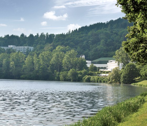 Rive du lac de barrage de Bitburg avec des arbres verts, un bâtiment en arrière-plan et un ciel dégagé., © TI Bitburger Land Rive du lac de barrage de Bitburg avec des arbres verts, un bâtiment en arrière-plan et un ciel dégagé., © TI Bitburger Land