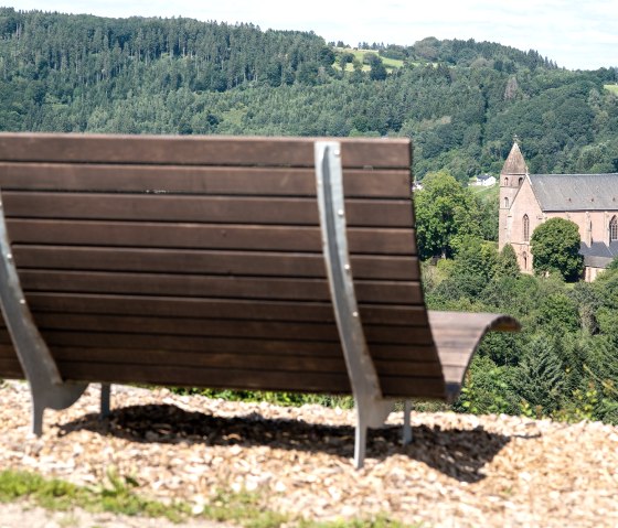 Eine Holzbank im Vordergrund bietet einen Ausblick auf eine Kirche, umgeben von grünen Bäumen und Hügeln. Eine Holzbank im Vordergrund bietet einen Ausblick auf eine Kirche, umgeben von grünen Bäumen und Hügeln.