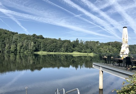 Het stuwmeer van Bitburg met rustig water, omgeven door bos. Een terras met tafels en parasol steekt in het beeld. Heldere lucht met dampsporen., © TI Bitburger Land Het stuwmeer van Bitburg met rustig water, omgeven door bos. Een terras met tafels en parasol steekt in het beeld. Heldere lucht met dampsporen., © TI Bitburger Land