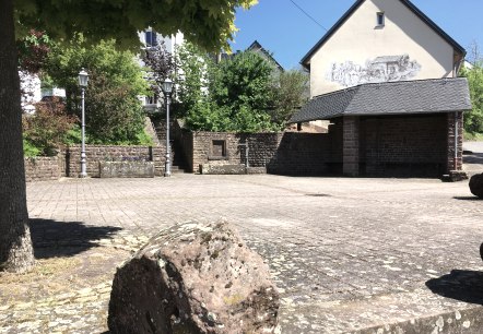 Een rustig dorpsplein met een geplaveide vloer, omringd door bomen en een gebouw met muurschilderingen. Lantaarns en planten sieren het plein., © Tourist-Information Bitburger Land Een rustig dorpsplein met een geplaveide vloer, omringd door bomen en een gebouw met muurschilderingen. Lantaarns en planten sieren het plein., © Tourist-Information Bitburger Land