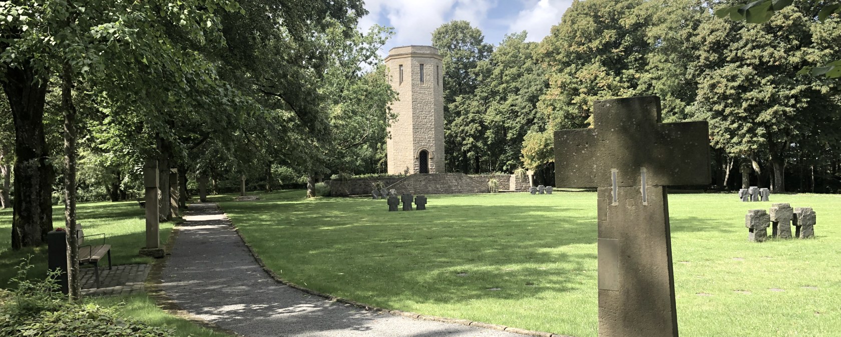 A cemetery with stone crosses, a tower in the background and a path lined with trees. The sky is partly cloudy. A cemetery with stone crosses, a tower in the background and a path lined with trees. The sky is partly cloudy.