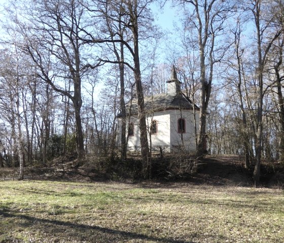 A small chapel stands in a sparse forest with bare trees. The sky is clear and blue., © Eifelverein Ortsgruppe Speicher A small chapel stands in a sparse forest with bare trees. The sky is clear and blue., © Eifelverein Ortsgruppe Speicher