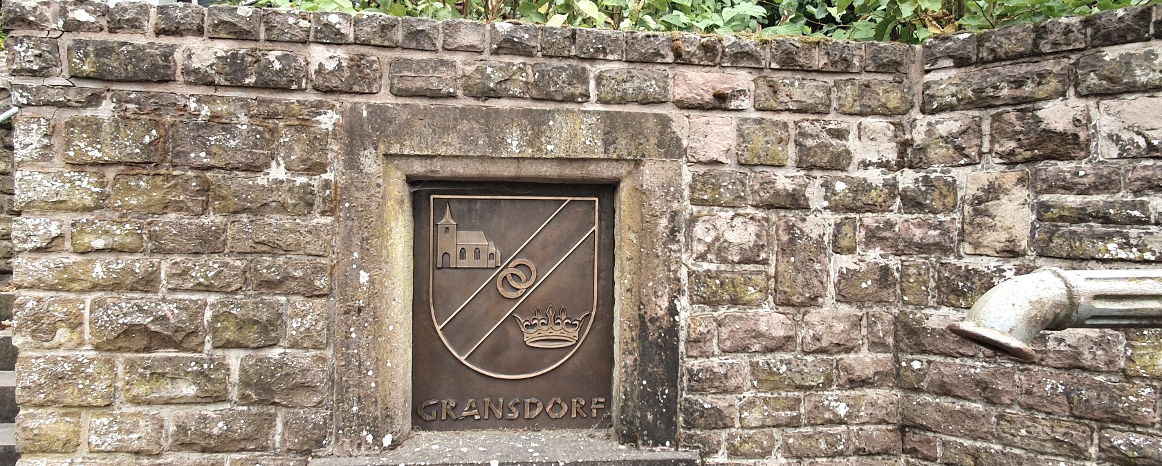 Mur de pierre avec un blason de Gransdorf représentant une église, deux anneaux et une couronne. A droite, on peut voir un tuyau d'évacuation., © TI Bitburger Land S.Wagner Mur de pierre avec un blason de Gransdorf représentant une église, deux anneaux et une couronne. A droite, on peut voir un tuyau d'évacuation., © TI Bitburger Land S.Wagner