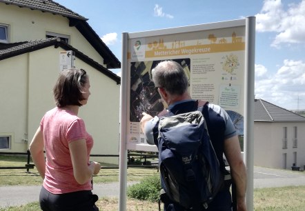 Two hikers stand in front of a hiking board with the inscription 'Metterich wayside crosses' and find out about the route. Houses can be seen in the background., © Tourist-Information Bitburger Land Two hikers stand in front of a hiking board with the inscription 'Metterich wayside crosses' and find out about the route. Houses can be seen in the background., © Tourist-Information Bitburger Land