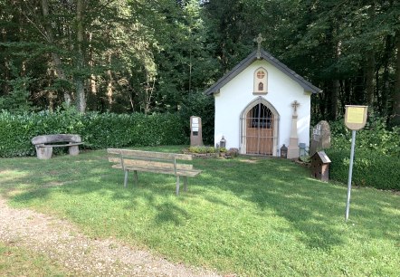 Eine kleine Kapelle im Wald mit einer Bank und einer Infotafel davor. Die Umgebung ist grün und friedlich., © TI Bitburger Land Eine kleine Kapelle im Wald mit einer Bank und einer Infotafel davor. Die Umgebung ist grün und friedlich., © TI Bitburger Land