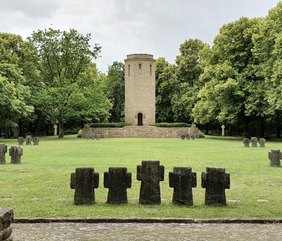 Ehrenfriedhof Kolmeshöhe in Bitburg: Ein zentraler Turm umgeben von grüner Wiese und mehreren Steinkreuzen, eingebettet in einen Wald. Ehrenfriedhof Kolmeshöhe in Bitburg: Ein zentraler Turm umgeben von grüner Wiese und mehreren Steinkreuzen, eingebettet in einen Wald.