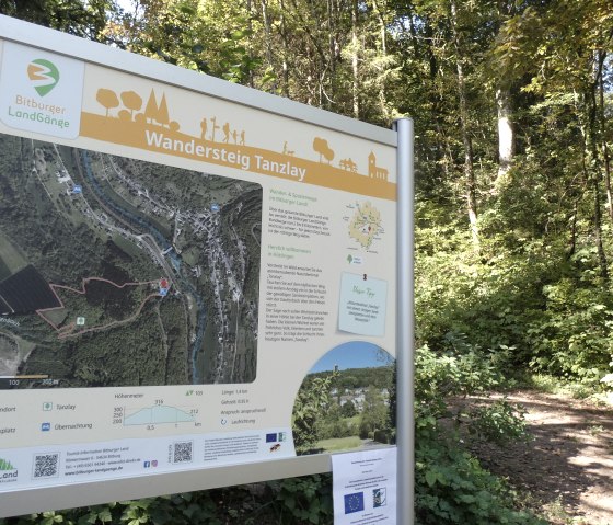 Startbord van de Tanzlay-wandelroute in het Bitburger Land, omgeven door dichte bossen en een wandelpad., © Tourist Information Bitburger Land Startbord van de Tanzlay-wandelroute in het Bitburger Land, omgeven door dichte bossen en een wandelpad., © Tourist Information Bitburger Land