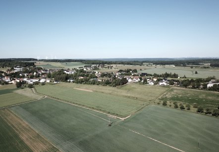 Luchtfoto van Idenheim: een landelijke nederzetting omringd door groene velden, met windturbines aan de horizon onder een heldere hemel., © Ingrid Penning Luchtfoto van Idenheim: een landelijke nederzetting omringd door groene velden, met windturbines aan de horizon onder een heldere hemel., © Ingrid Penning