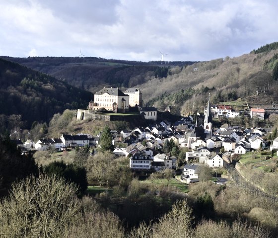 Schloss Malberg thront über einem malerischen Dorf in einer hügeligen Landschaft, umgeben von Wäldern und Windrädern im Hintergrund., © TI Bitburger Land Monika Mayer Schloss Malberg thront über einem malerischen Dorf in einer hügeligen Landschaft, umgeben von Wäldern und Windrädern im Hintergrund., © TI Bitburger Land Monika Mayer