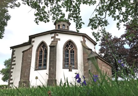La chapelle Frohnert avec ses fenêtres gothiques, entourée d'arbres et de fleurs au premier plan, photographiée depuis une perspective basse., © TI Bitburger Land La chapelle Frohnert avec ses fenêtres gothiques, entourée d'arbres et de fleurs au premier plan, photographiée depuis une perspective basse., © TI Bitburger Land