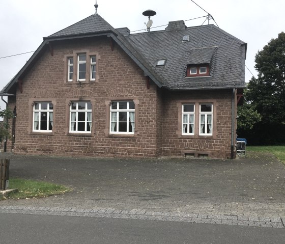 Een oud bakstenen gebouw met een leistenen dak en witte ramen staat naast een straat in Wilsecker. Een boom en gazon zijn zichtbaar op de achtergrond., © Tourist-Info Bitburger Land M.Mayer Een oud bakstenen gebouw met een leistenen dak en witte ramen staat naast een straat in Wilsecker. Een boom en gazon zijn zichtbaar op de achtergrond., © Tourist-Info Bitburger Land M.Mayer