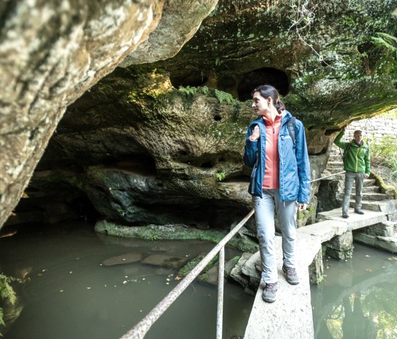 Twee mensen op een smalle loopbrug over een vijver, omgeven door rotsen en planten. De ene persoon draagt een blauwe jas, de andere een groene., © Eifel Tourismus, D. Ketz Twee mensen op een smalle loopbrug over een vijver, omgeven door rotsen en planten. De ene persoon draagt een blauwe jas, de andere een groene., © Eifel Tourismus, D. Ketz