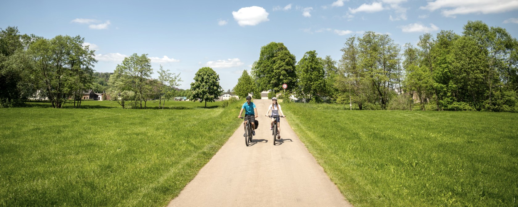 Zwei Radfahrer fahren auf einem asphaltierten Weg durch eine grüne Landschaft. Der Himmel ist blau mit einigen Wolken., © Eifel Tourismus GmbH, Dominik Ketz Zwei Radfahrer fahren auf einem asphaltierten Weg durch eine grüne Landschaft. Der Himmel ist blau mit einigen Wolken., © Eifel Tourismus GmbH, Dominik Ketz