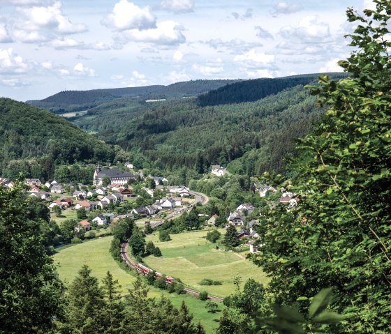 Vue panoramique d'un village au milieu de collines et de forêts verdoyantes. Un train circule sur une voie ferrée à travers le paysage., © TI Bitburger Land Vue panoramique d'un village au milieu de collines et de forêts verdoyantes. Un train circule sur une voie ferrée à travers le paysage., © TI Bitburger Land