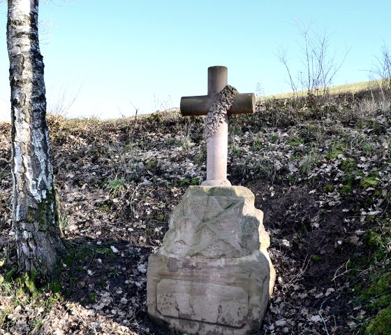 Une croix de chemin en pierre recouverte de lierre se trouve entre deux arbres sur une colline couverte de végétation., © TI Bitburger Land Une croix de chemin en pierre recouverte de lierre se trouve entre deux arbres sur une colline couverte de végétation., © TI Bitburger Land