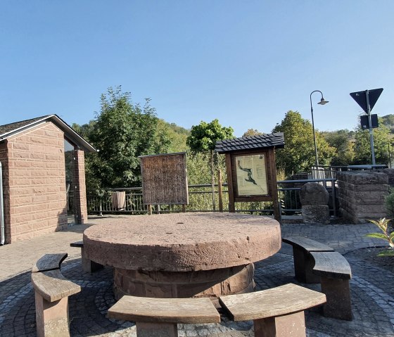 Village square in Brecht with round bench, information boards and small building. Surrounded by trees and a road in the background., © TI Bitburger Land, Steffi Wagner Village square in Brecht with round bench, information boards and small building. Surrounded by trees and a road in the background., © TI Bitburger Land, Steffi Wagner