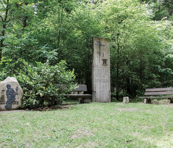 Aire de repos dans la forêt avec deux bancs en bois, un poteau en bois avec une inscription et une pierre avec un relief. Entouré d'arbres et de buissons verts., © TI Bitburger Land Aire de repos dans la forêt avec deux bancs en bois, un poteau en bois avec une inscription et une pierre avec un relief. Entouré d'arbres et de buissons verts., © TI Bitburger Land