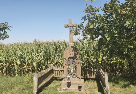 Een stenen kruis met een Jezusfiguur staat voor een maïsveld, omringd door bomen en een houten hek. De lucht is helder en blauw., © TI Bitburger Land Een stenen kruis met een Jezusfiguur staat voor een maïsveld, omringd door bomen en een houten hek. De lucht is helder en blauw., © TI Bitburger Land